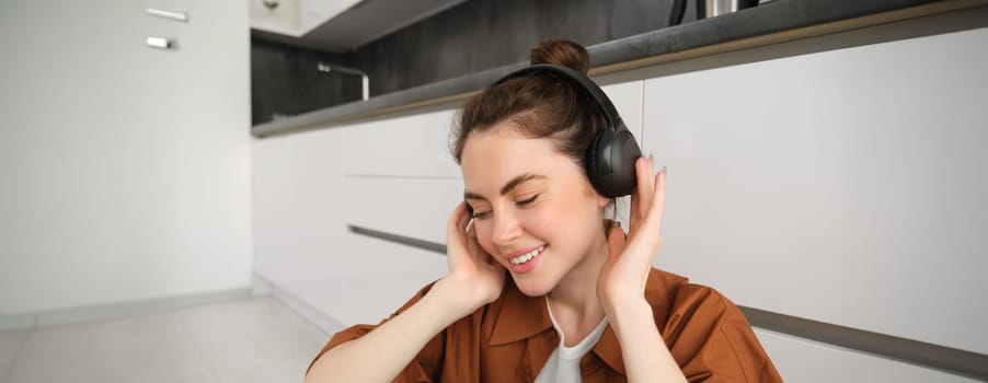 Cute brunette woman on kitchen floor, wears headphones, listens music and smiles, enjoys sound quality.