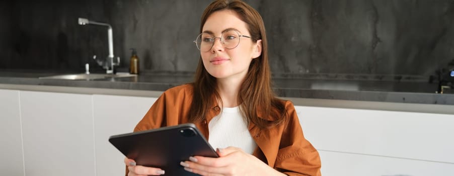 Image of beautiful young woman in glasses, sitting in her kitchen with digital tablet, watching videos online, reading, working on remote, studying remotely.