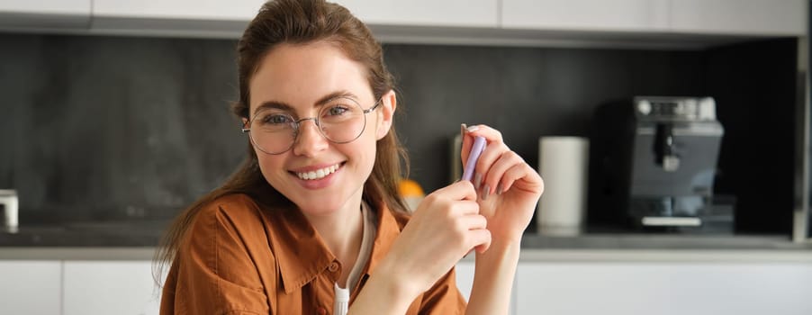 Portrait of beautiful young woman, entrepreneur in glasses, has own business from home, sitting in kitchen with pen, smiling and looking confident at camera.