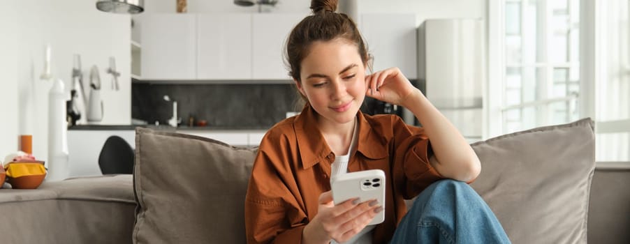 Portrait of young modern woman reading on mobile phone, scrolling social media app on smartphone, sitting on couch in living room in casual clothes.