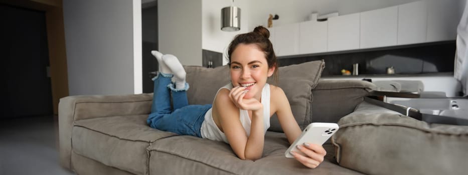 Portrait of cute, happy young woman lying on couch, holding mobile phone, using smartphone while relaxing at home in living room, concept of lifestyle and people leisure.