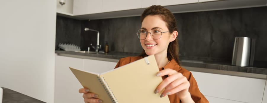 Portrait of beautiful woman in glasses, college girl smiling, sitting in kitchen with notebook.