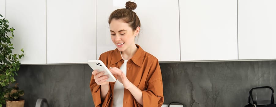 Image of beautiful young female model, posing with smartphone in the kitchen, looking at mobile screen and smiling while reading a message on phone.