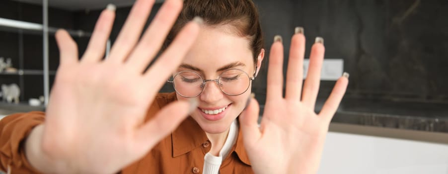 Portrait of cute and romantic young woman in glasses, covers her face, extends hands to block camera, asks to stop taking pictures.