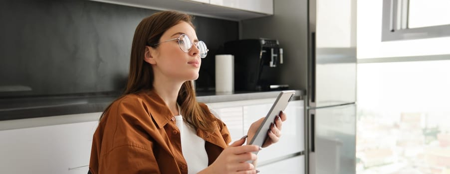 Portrait of young woman sitting at home in kitchen, holding digital tablet, wearing glasses, looking thoughtful, working on project, studying.