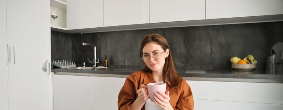 Portrait of happy young woman in glasses, sitting at home in kitchen, holding cup of coffee, drinking favourite tea, relaxing indoors on cold autumn day.