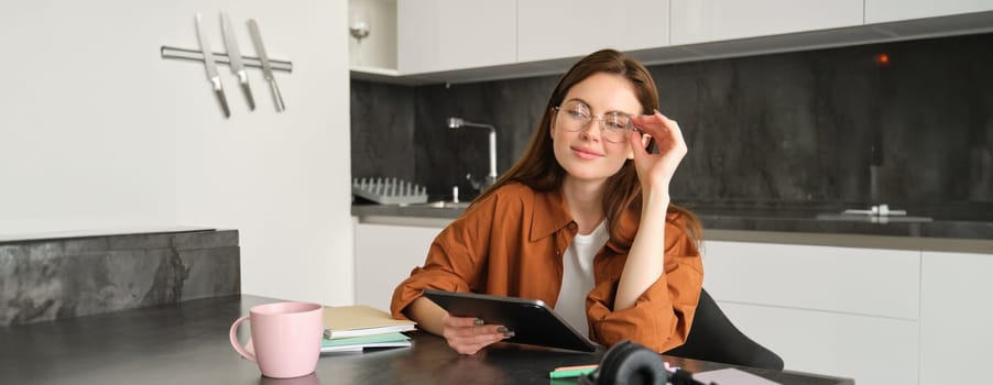Portrait of young student, woman studying at home, working remotely, setup workplace in her kitchen, sitting on chair with digital tablet, reading in glasses.