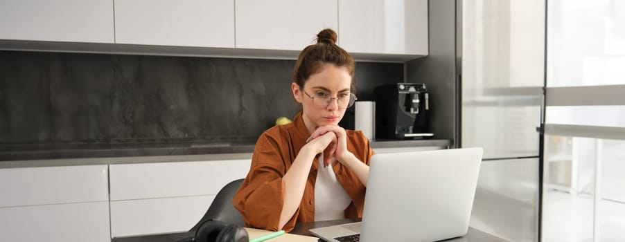Freelancing, work from home concept. Young woman sitting with laptop in kitchen, freelancer digital nomad doing her job on remote, using computer, looking thoughtful, thinking.
