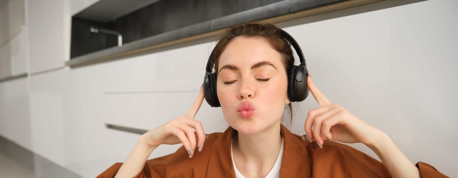 Close up of cute young woman, listens music in wireless headphones, makes kissing puckered lips and closes eyes, poses in earphones.