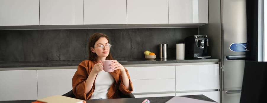 Portrait of young woman enjoying cup of coffee in peace, sitting at home, holding mug with herbal tea, wearing glasses, relaxing at in kitchen.