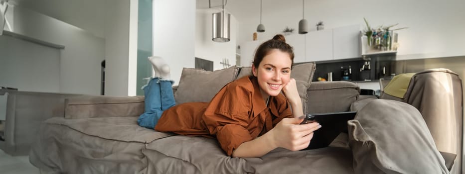 Portrait of cute young woman relaxing at home, watching movie on digital tablet or reading e-book, using gadget while lying on couch in living room, smiling at camera.