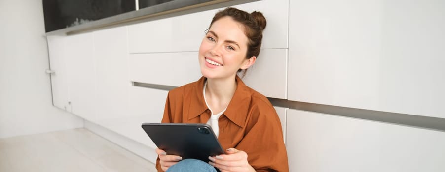 Young entrepreneurs. Smiling cute woman with digital tablet, sitting with gadget on kitchen floor and looking happy, studying remotely, reading on her device.