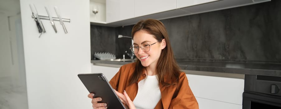 Portrait of girl in glasses, using digital tablet in kitchen, working from home, touches screen and smiling.