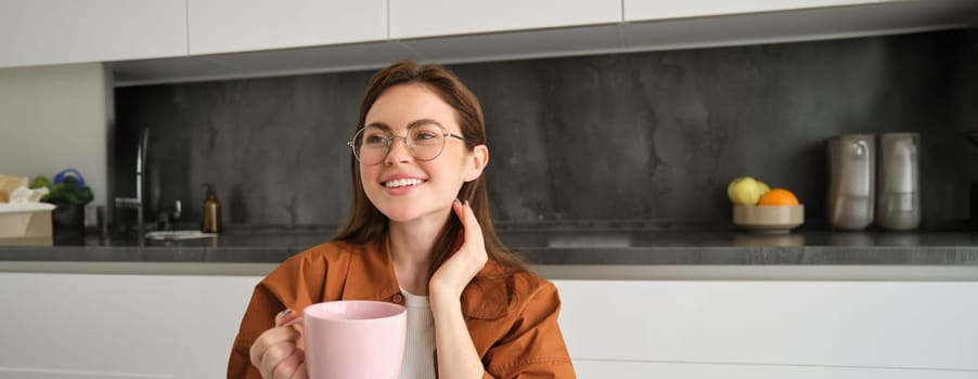 Portrait of beautiful young woman in glasses, smiling, resting at home, holding mug with coffee, drinking tea.