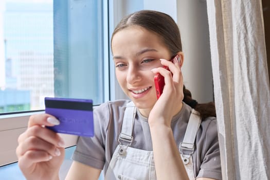 Young teenage student girl with credit bank card talking on cell phone, at home. Online payment for goods and services, shopping, credit cards for teenagers concept