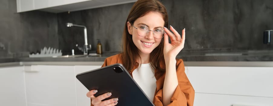 Portrait of beautiful young female model in glasses, holding digital tablet, wearing glasses, reading e-book, posing in kitchen at home.