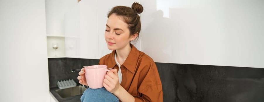 Portrait of beautiful young woman enjoying her morning cup of aromatic coffee, drinking tea while sitting in kitchen and contemplating sunny day.