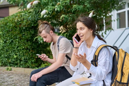 Teenage high school students, guy and girl, sitting on bench with backpacks, near academic building, using smartphones. Adolescence, youth, education, lifestyle, technology concept