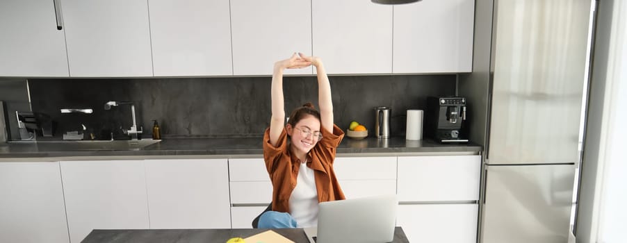Productivity and lifestyle concept. Beautiful young woman finishes working, stretching hands with satisfied expression, sitting in kitchen with laptop and documents, has remote workplace at home.