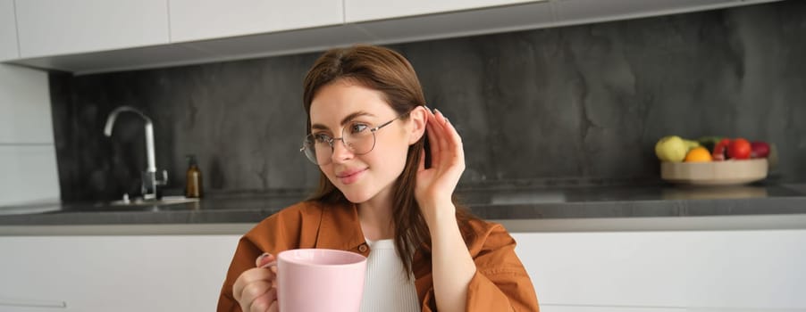 Portrait of lovely, tender young woman in glasses, sits at home with cup of tea. Girl in kitchen, enjoys drinking aromatic coffee.