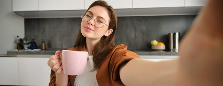 Portrait of beautiful young woman in glasses, takes selfie with cup of tea, sitting in kitchen, holding smartphone.