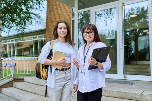 Female teacher, mentor, coach and teenage girl student with backpack together, looking at camera outdoor, school building background. Adolescence, education, knowledge, communication