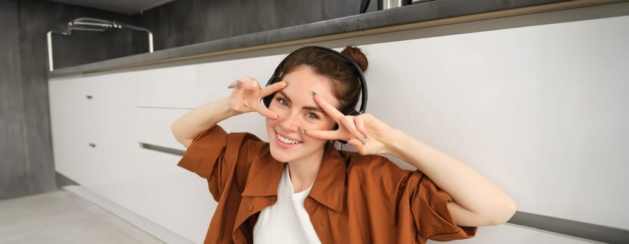 Happy young woman in headphones, listens music, enjoys sound quality, shows v-sign.