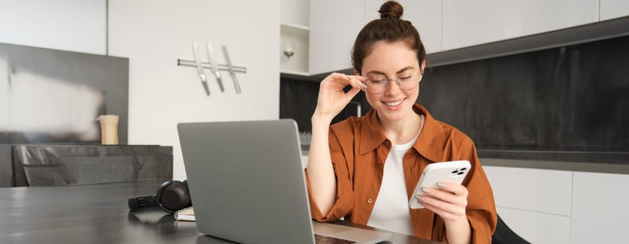 Portrait of young self-employed woman, replying to customers on smartphone, using laptop to check orders, sitting at home.