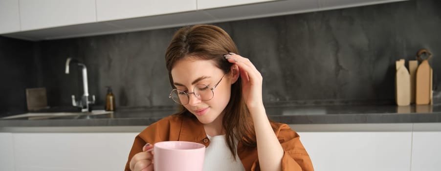 Close up portrait of cute and tender young woman in glasses, sitting in kitchen, drinking tea, holding cup, tucks her hair behind ear and smiles.