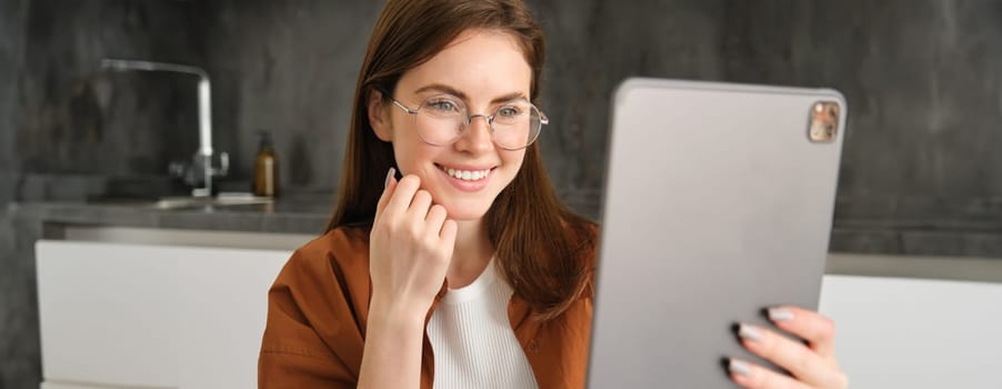 Portrait of beautiful young woman in glasses, looking at digital tablet. Businesswoman works on remote, freelancer reading on gadget, sitting in kitchen.