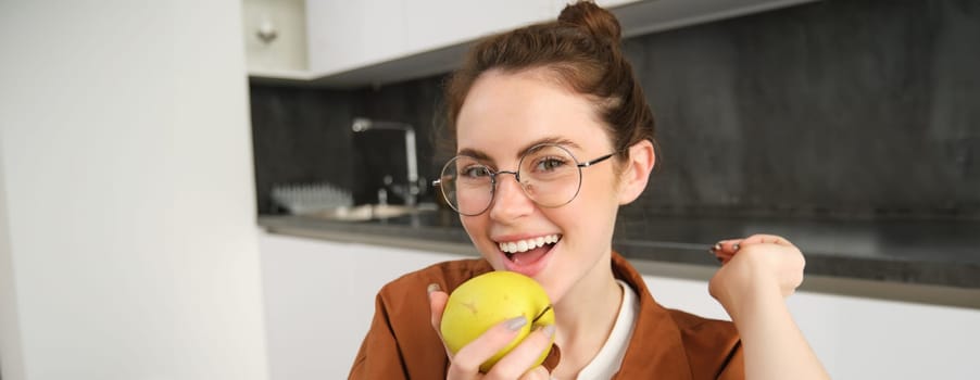 Close-up portrait of brunette woman at home, wearing glasses, eating apple in the kitchen and smiling, biting fruit.