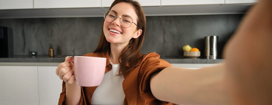 Carefree girl in glasses, takes selfie with smartphone, smiling and looking happy, enjoying hot drink in kitchen at home.