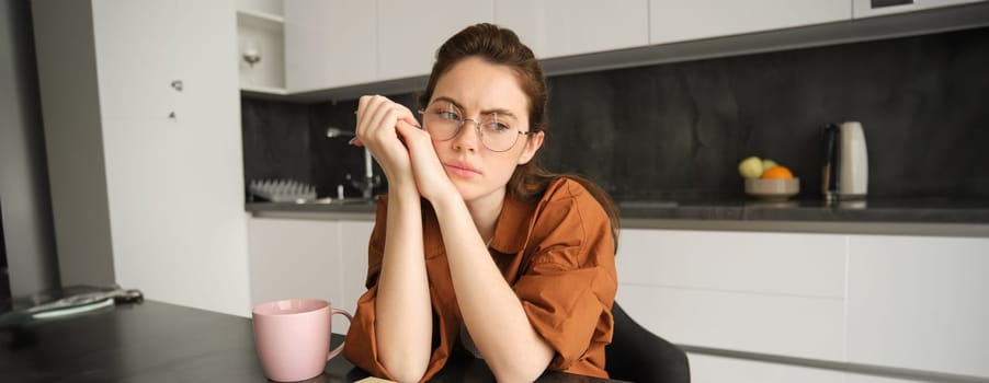 Portrait of young woman with troubled face, wearing glasses, sitting in kitchen with work materials on table, looking aside concerned, tired of working, studying at home.