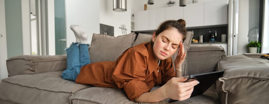 Portrait of young woman feeling unwell, lying on couch with digital tablet, touching head and frowning from painful migraine, has headache.