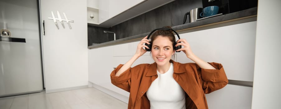 Carefree woman listens to music at home, sits on floor, puts on wireless headphones, smiles and looks happy.