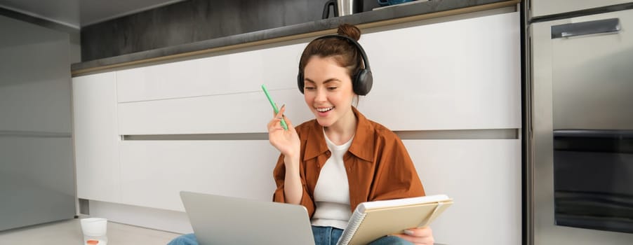 Woman studies, does homework at home, connects to online lesson, sits on floor with laptop, writes in notebook, works in headphones.