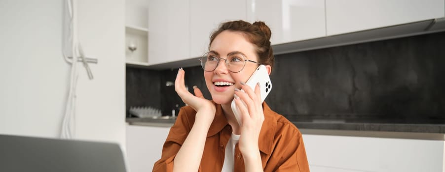 Portrait of young businesswoman, self-employed lady working from home, making phone calls to clients and smiling, using laptop in kitchen.