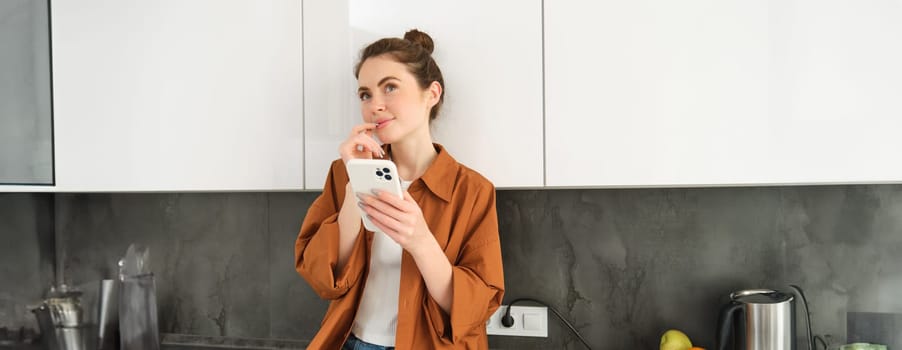 Portrait of woman online shopping using smartphone app, sitting in kitchen with thinking face, holding mobile phone.