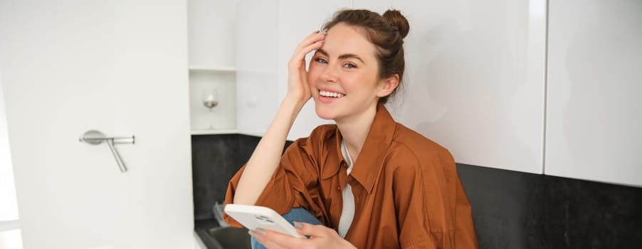 Portrait of young woman with mobile phone, browsing social media, online shopping, sitting on kitchen counter, smiling and looking happy.