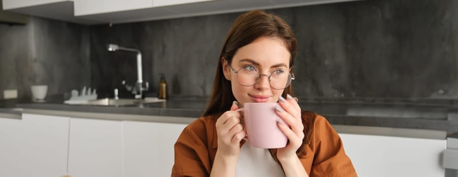 Portrait of beautiful young woman in glasses, drinking coffee, holding cup and enjoying her tea in kitchen.