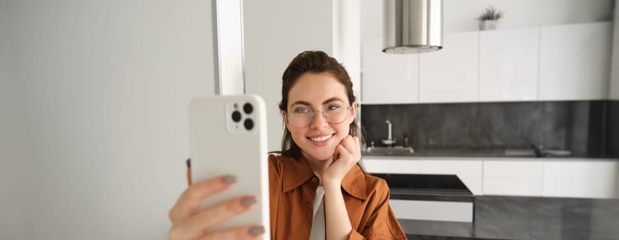 Beautiful young woman in glasses taking selfie in her flat, using smartphone, posing for photo, standing with mobile phone and smiling.