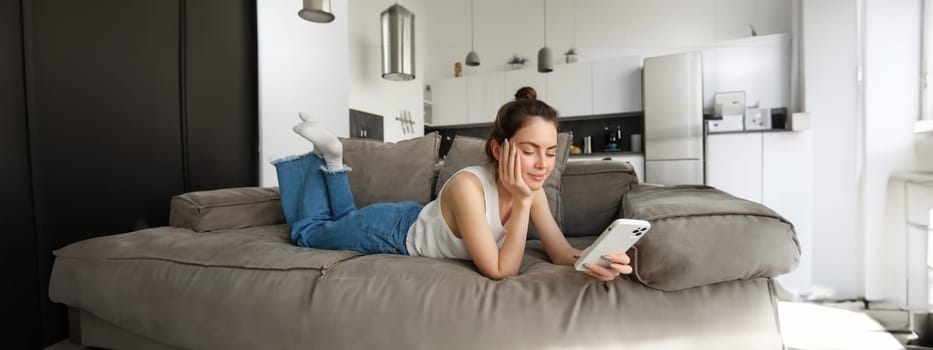 Portrait of smiling, happy young woman resting at home, resting on couch with smartphone, using mobile phone application.