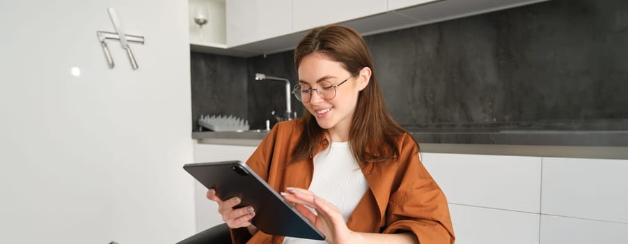Young woman at home, reading in glasses on digital tablet and smiling, sitting in kitchen.