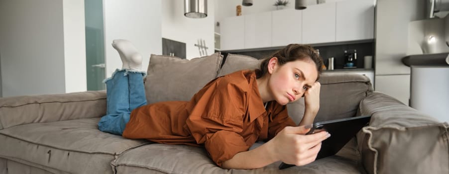 Portrait of woman lying on couch upset, using digital tablet, looking disappointed and sulking, spending time at home.