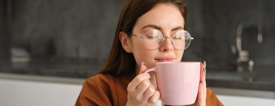 Close up portrait of beautiful brunette woman in glasses, smells her coffee, holds cup, drinks tea at home in kitchen, smiling pleased.