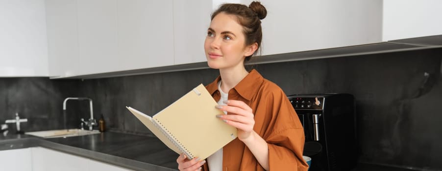 Portrait of gorgeous young woman reading, student standing in the kitchen with planner, doing homework, revising for exam.
