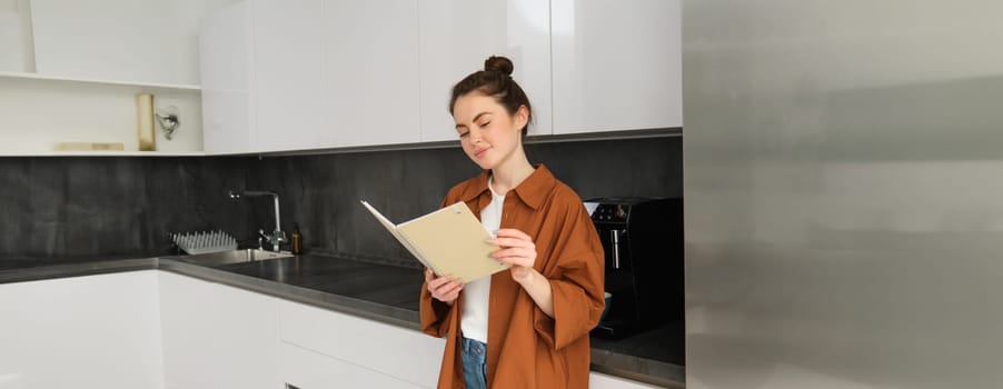 Portrait of beautiful woman standing in the kitchen with notebook, reading recipes, flipping pages and smiling.