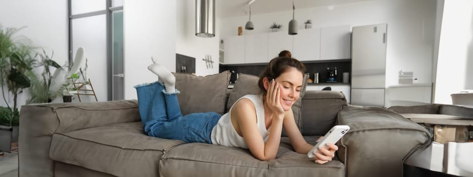 Portrait of cute young woman with smartphone, lying on couch and resting at home, scrolling social media feed, online shopping, reading something on mobile phone.