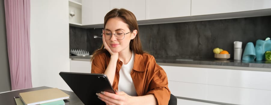 Portrait of young woman, freelancer sitting in kitchen, reading on digital tablet, working from home, studying online, has remote education.