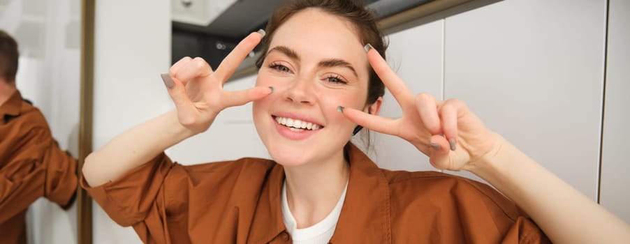 Close up portrait of happy, beautiful girl listens to music in wireless headphones, shows peace, v-sign gestures and smiles, enjoys songs in favourite playlist.